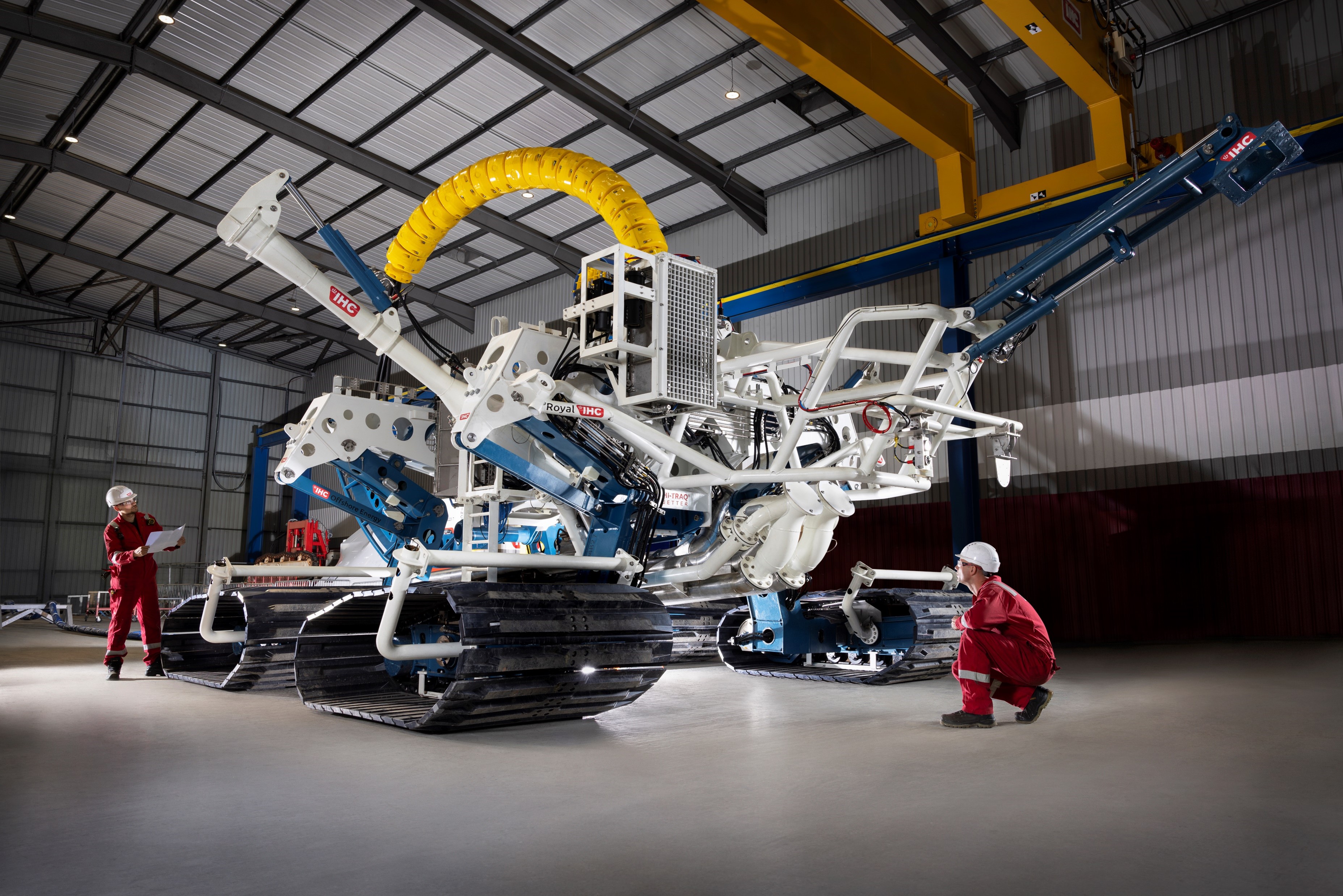 Four-tracked blue and white subsea trencher in a large, modern factory bay. Two workers in red overalls and white hard hats stand either side.