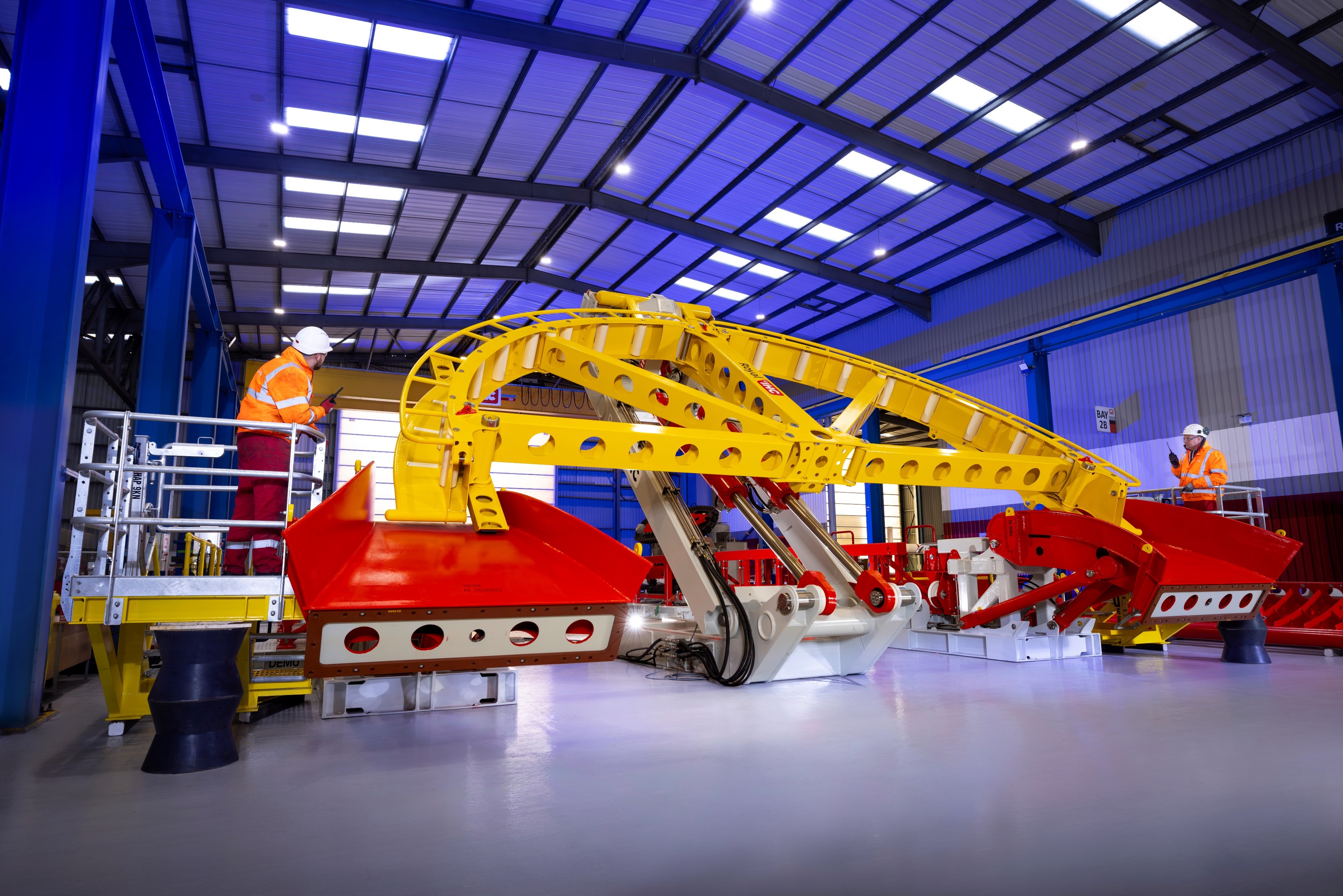 In a large factory bay, the yellow quadrant is tipping forward. Two workers dressed in orange hi-vis jackets and white hard hats stand either side