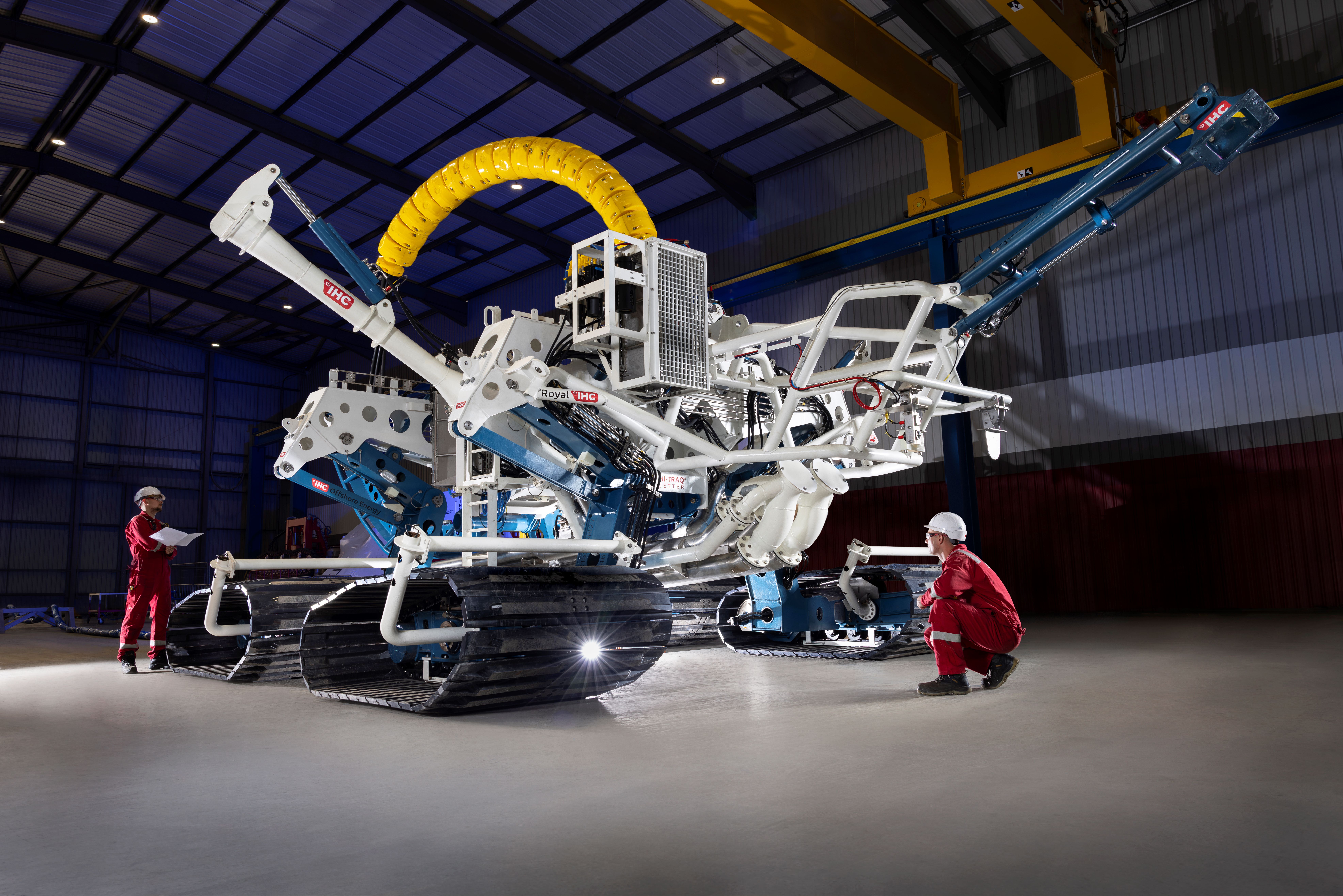 Four-tracked white subsea trencher in a large, clean factory bay. Two workers either side of the trencher in red overalls and white hard hats.