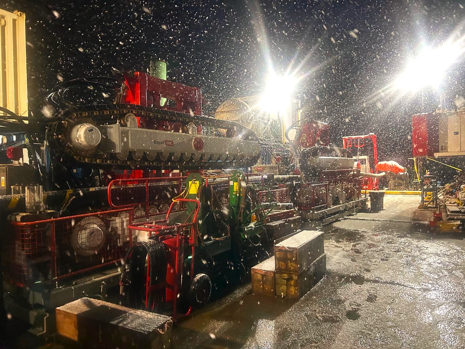 Two red and white cable tensioners in operation on a vessel deck at night, with snow falling.