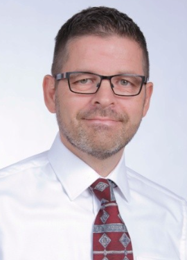 Headshot photo of Ed wearing a white shirt and red tie. He is smiling in front of a white background.