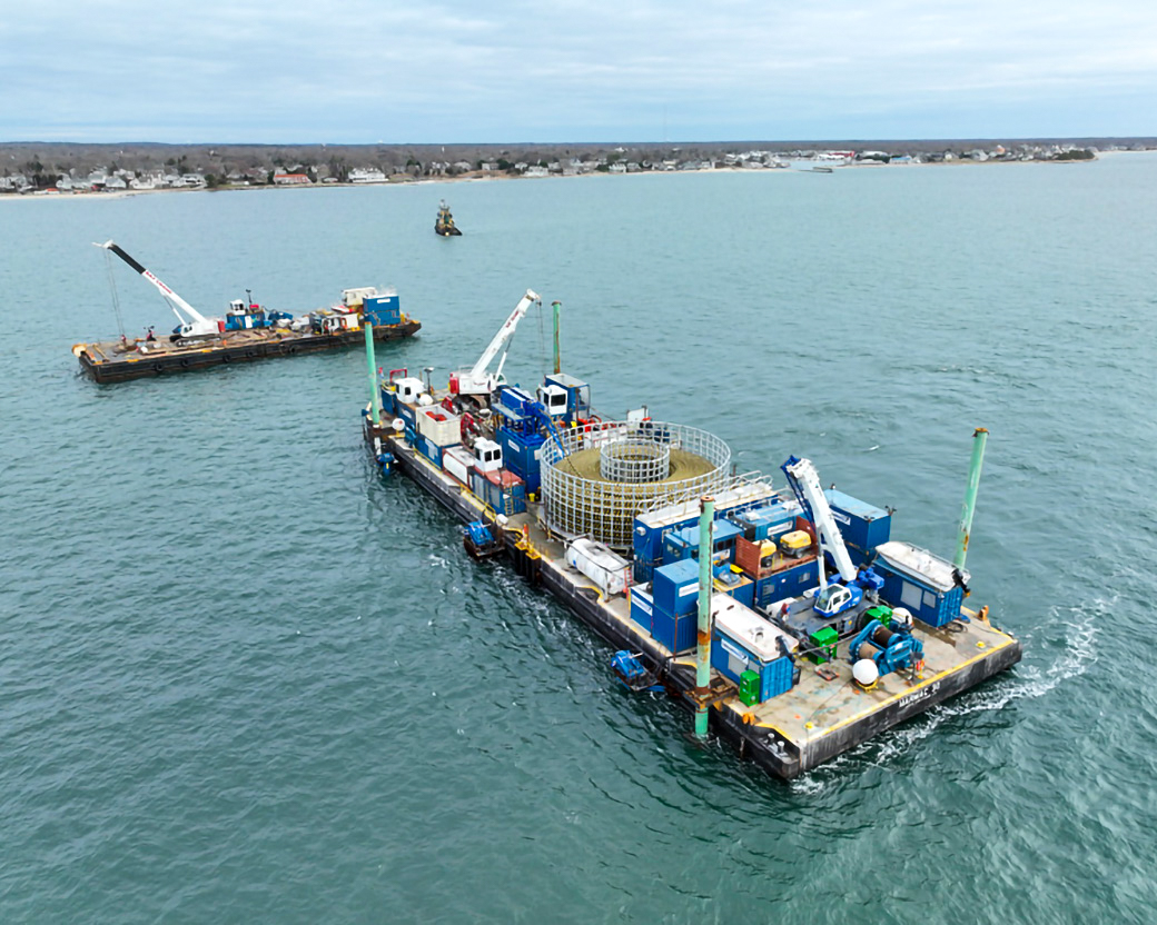Aerial view of a cable lay barge on calm blue waters, with the coast visible in the background.