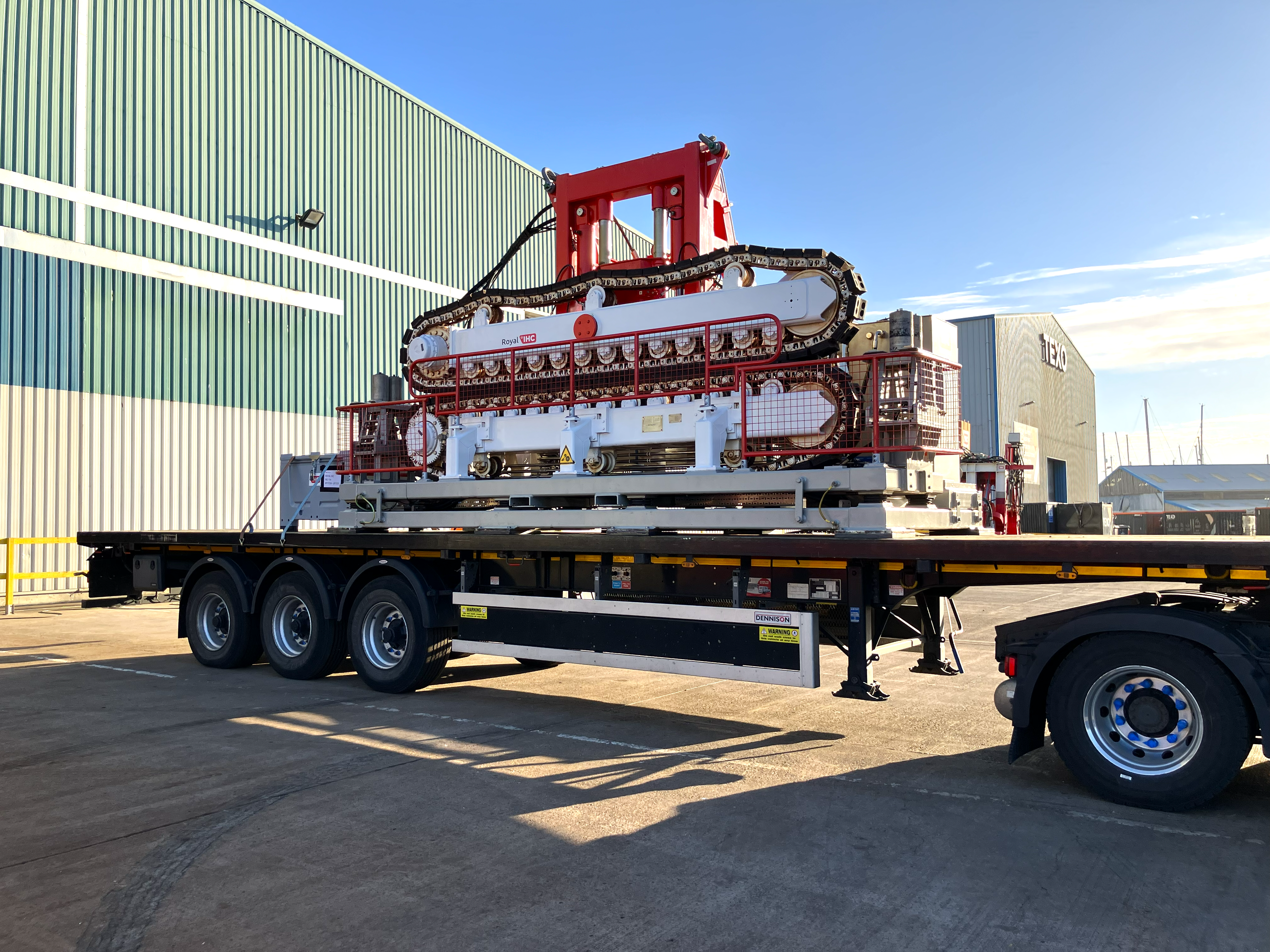 Red and white cable tensioner loaded on the back of an open top tuck in front of a factory, with blue skies behind.