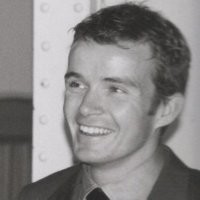 Black and white photo of man with short dark hair and wearing a collared shirt. Smiling and looking to the left..