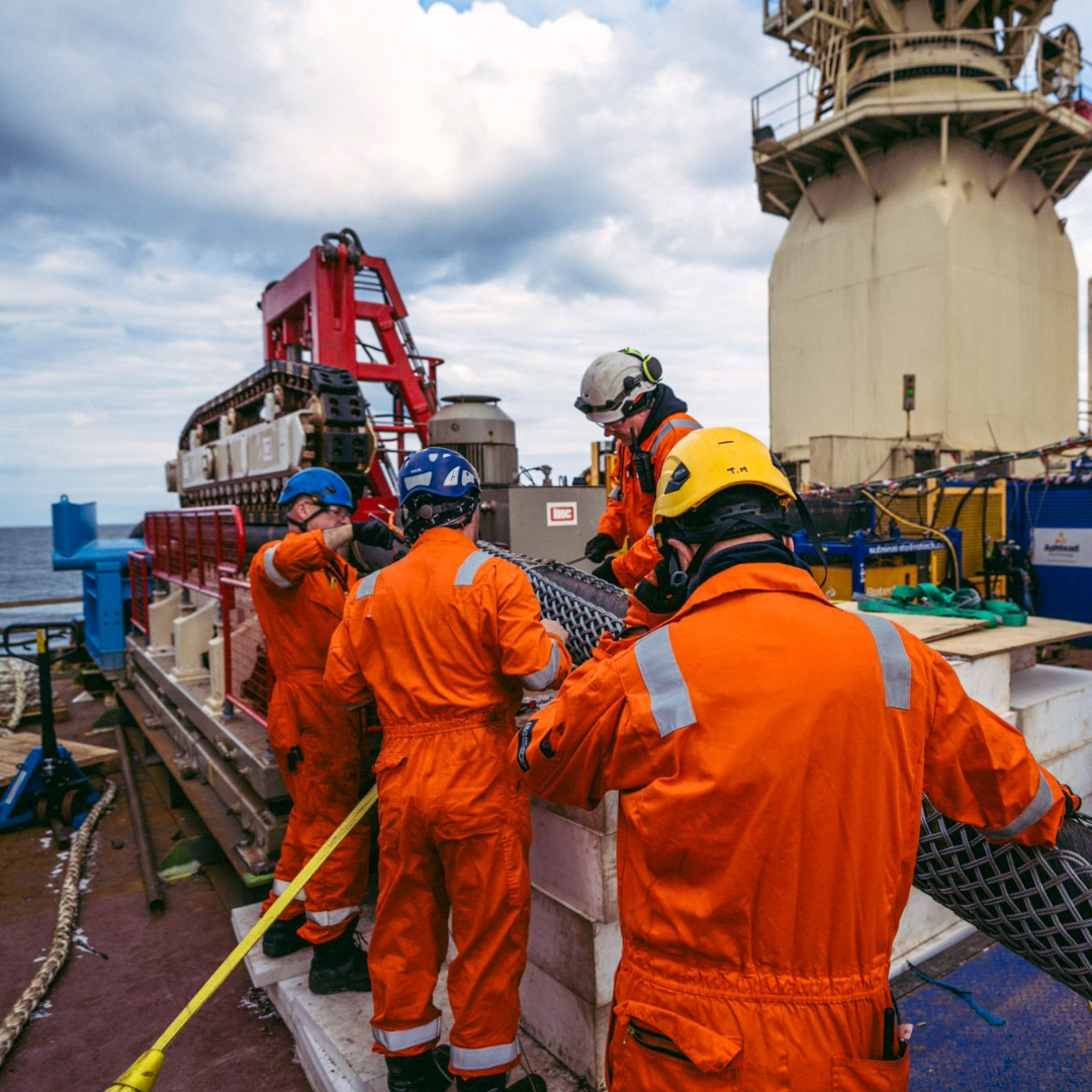 Four workers wearing hard hats and orange overalls onboard a vessel, working with the red and white cable tensioner. Cloudy sky in the background.